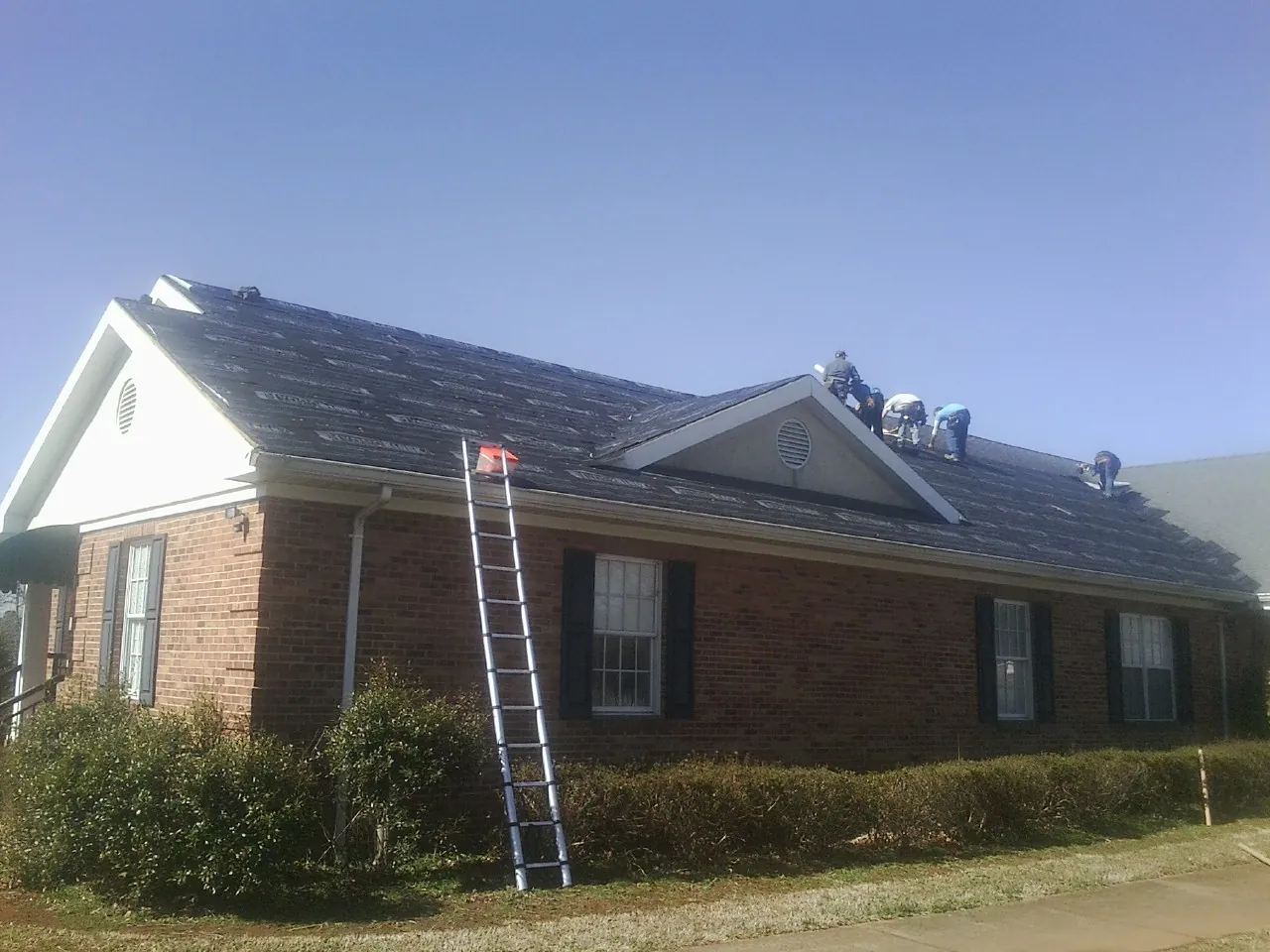 Crew tearing shingles off a brick ranch roof with a ladder set — Emergency Roof Repair work by our crew in Stokesdale