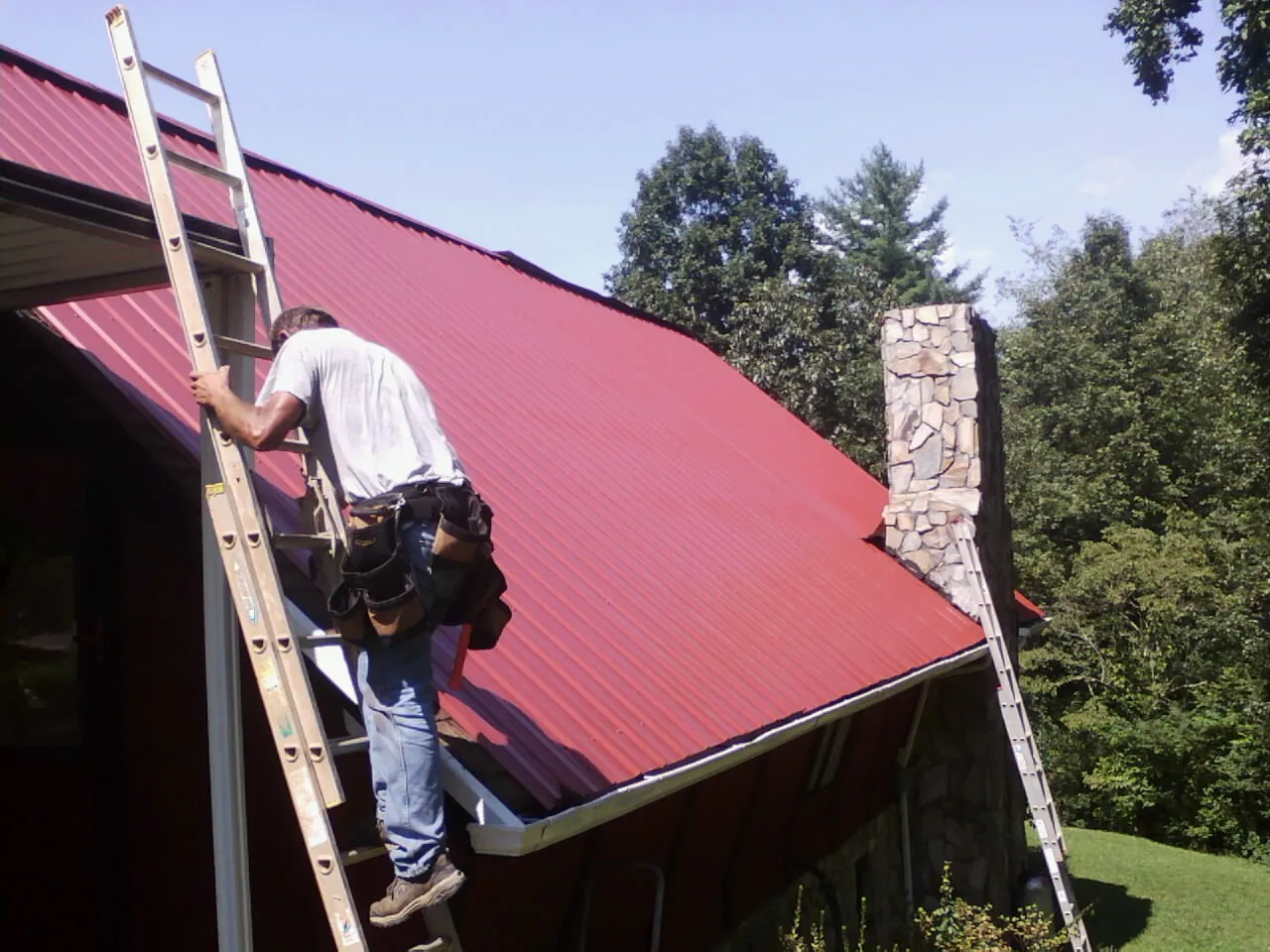 Roofer on a ladder beside a red metal roof and stone chimney — Soffit & Fascia Repair by Roofing Brothers in Stokesdale